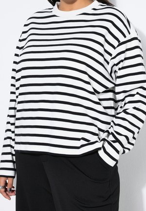Personne portant une chemise blanche à manches longues avec des rayures horizontales noires et un pantalon noir, une main dans la poche et l'autre détendue sur le côté.
