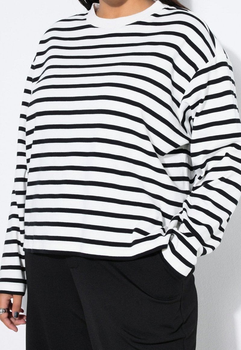 Personne portant une chemise blanche à manches longues avec des rayures horizontales noires et un pantalon noir, une main dans la poche et l'autre détendue sur le côté.