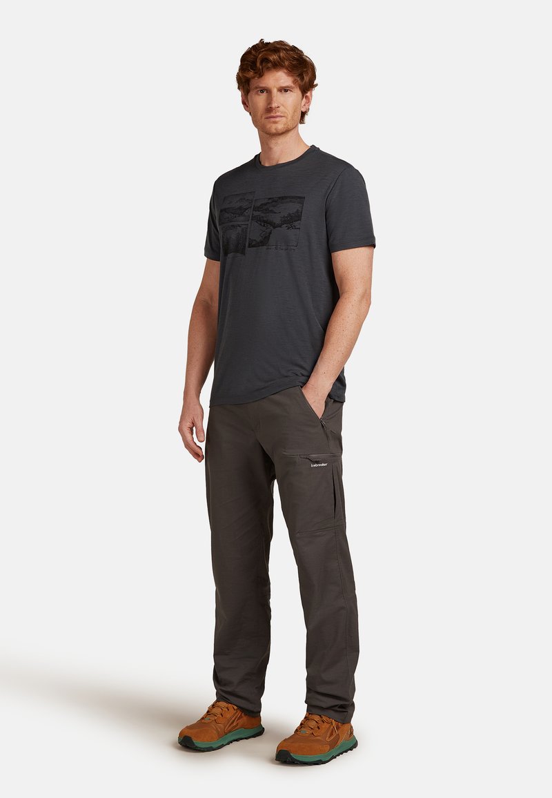 Hombre de pie con camiseta gráfica de color gris oscuro, pantalones de cargo oscuros y zapatos de senderismo marrones, frente a un fondo blanco.