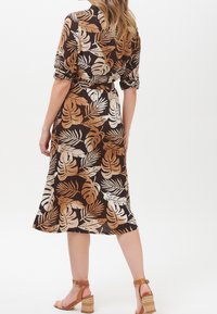 Donna in piedi con le spalle girate, che indossa un vestito marrone con motivi di foglie tropicali color sabbia e beige e sandali con tacco quadrato color sabbia.