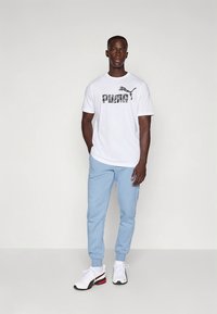 Biała koszulka z wyrazistym szarym logo „PUMA”, jasnoniebieskie dresy oraz czarno-białe obuwie z czerwonymi akcentami. Prosty design, wygodne dopasowanie.