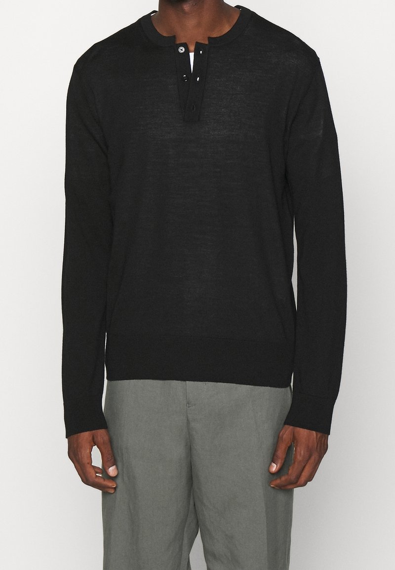 Homme portant un t-shirt henley noir à manches longues avec trois boutons et un pantalon gris, debout devant un fond uni.