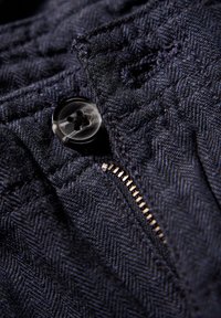 Marinblått strukturerat tyg med en fiskbensmönstring, med en svart plastknapp och en metallisk dragkedjedetalj.