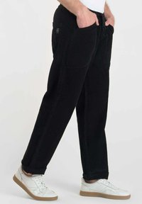 Persoon in zwarte ribfluwelen broek met voorzakken en witte sneakers met bruine zolen tegen een effen achtergrond.