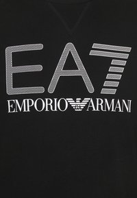 Svart tyg med vit grafisk text "EA7" ovanför "Emporio Armani"-logotypen som har en örn med ränder mellan orden.