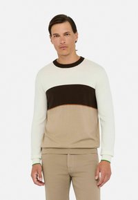 Man in beige broek en een lange mouwen trui met witte, donkerbruine en lichtbruine horizontale strepen, staand voor een effen achtergrond.