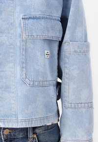 Veste en denim bleu clair avec une coupe légèrement surdimensionnée, dotée de deux poches avant, de coutures visibles et d'une petite étiquette de marque.