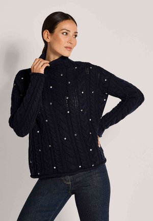 Frau trägt schwarzen Kabelstrickpullover mit kleinen weißen Perlen, posiert mit einer Hand auf der Hüfte und der anderen in der Nähe des Halses.