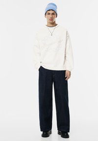 Giovane uomo che indossa un berretto light blue, una felpa crema con motivo a stelle, jeans larghi scuri, scarpe nere e una collana d'argento, in piedi contro uno sfondo bianco.