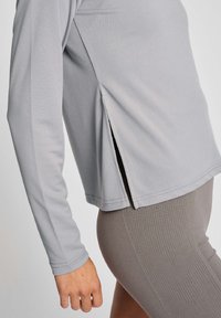 Hellgraues langärmliges Oberteil mit seitlichen Schlitzen, hergestellt aus atmungsaktivem Stoff. Verfügt über nahtlose Bündchen und eine lockere Passform. Kombiniert mit strukturierten grauen Leggings.