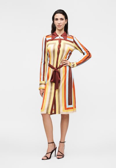 Γυναίκα με σκούρα μαλλιά που φορά φόρεμα μέχρι το γόνατο με μακριά μανίκια, κουμπιά, πολύχρωμες κάθετες και διαγώνιες ρίγες και δεμένη ζώνη στη μέση.