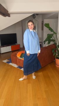Γυναίκα με μπλε μπλούζα και μακριά σκούρα μπλε φούστα που χαμογελά μέσα σε ένα μοντέρνο σαλόνι με ξύλινο πάτωμα και μεγάλο πράσινο φυτό.