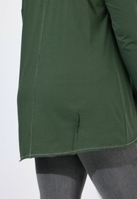 Haut vert à manches longues en tissu extensible avec un ourlet arrondi et des fentes latérales ; présente des coutures visibles le long des seams et une texture lisse.