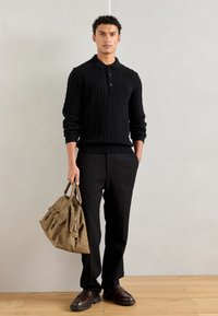 Czarny sweter w splot warkoczowy z kołnierzykiem, czarne spodnie, brązowe skórzane buty oraz beżowa torba z wieloma kieszeniami i zamkiem.