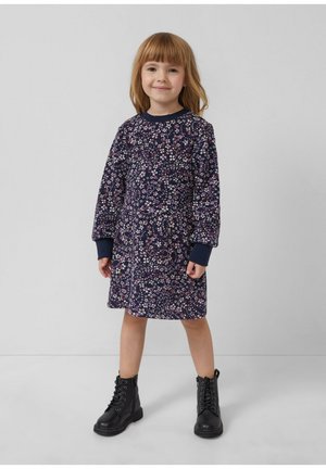 Junges Mädchen mit hellbraunen Haaren, das ein marineblaues, blumiges Langarmkleid und schwarze Schnürstiefel trägt, steht vor einem schlichten grauen Hintergrund.
