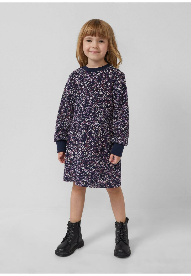 Junges Mädchen mit hellbraunen Haaren, das ein marineblaues, blumiges Langarmkleid und schwarze Schnürstiefel trägt, steht vor einem schlichten grauen Hintergrund.