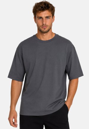 Jonge man met kort bruin haar en getrimde baard draagt een losvallend donkergrijs T-shirt en zwarte broek, staand tegen een effen lichte achtergrond.