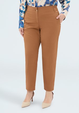 Kvinde iført brune højtaljede taperbukser kombineret med beige spidse hæle og en blå blomstret bluse, hænderne i lommerne.