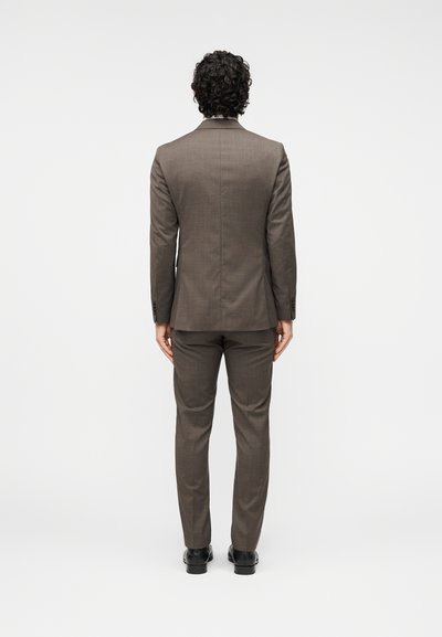 Καφέ κοστούμι με στενή εφαρμογή, από υφή ύφασμα, με γ ιακάδες notch και μονό σκίσιμο στο σακάκι, συνδυασμένο με αντίστοιχο παντελόνι και μαύρα παπούτσια.