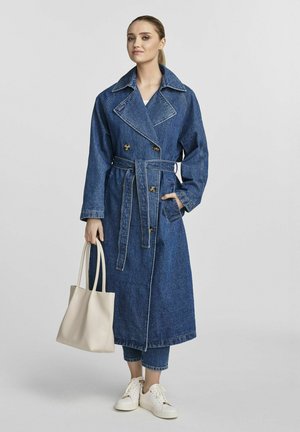 Denimowy trencz o podwójnym zapięciu, szerokim kołnierzu, pasku w talii i bocznych kieszeniach. Noszony z beżową torbą i białymi trampkami.