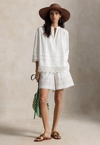 Γυναίκα που φοράει ψάθινο καπέλο με πλατύ γείσο, λευκή μπλούζα με δαντέλα και σορτς, καφέ σανδάλια, και κρατάει μια πράσινη τσάντα με ρίγες.