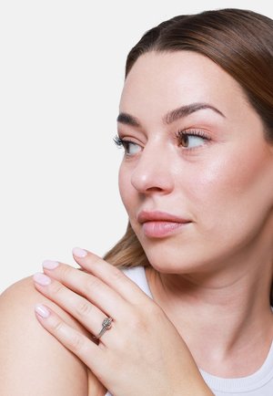 Kobieta o brązowych włosach patrzy w bok, opierając rękę z srebrnym pierścieniem w kształcie węzła na ramieniu, nosi białą koszulkę bez rękawów, neutralne tło.