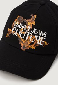 Čierny baseballový klobúk s živou zlatou a oranžovou kvetinovou potlačou, s textom "VERSACE JEANS COUTURE" v bielej farbe integrovaným do dizajnu.