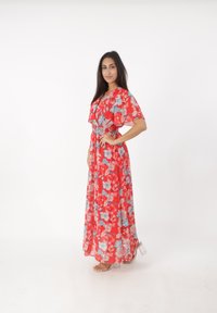 Robe maxi rouge à fleurs avec de courtes manches volantes, taille élastique et superposition transparente. Présente des motifs de fleurs bleues et blanches. Talons transparents.