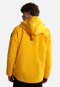 Yellow hooded jacket, made of smooth fabric, features a drawstring and a logo patch on the left shoulder. The fit is slightly oversized.