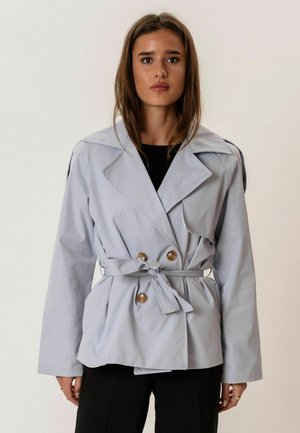 Nő világosszürke, kétsoros gombolású ballonkabátot visel, amelyet deréknál övvel kötött meg, és egyszínű háttér előtt áll.