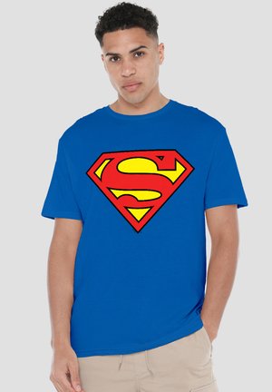 Young man with short curly hair wearing a blue t-shirt featuring a large red and yellow Superman logo, standing with one hand in pocket.