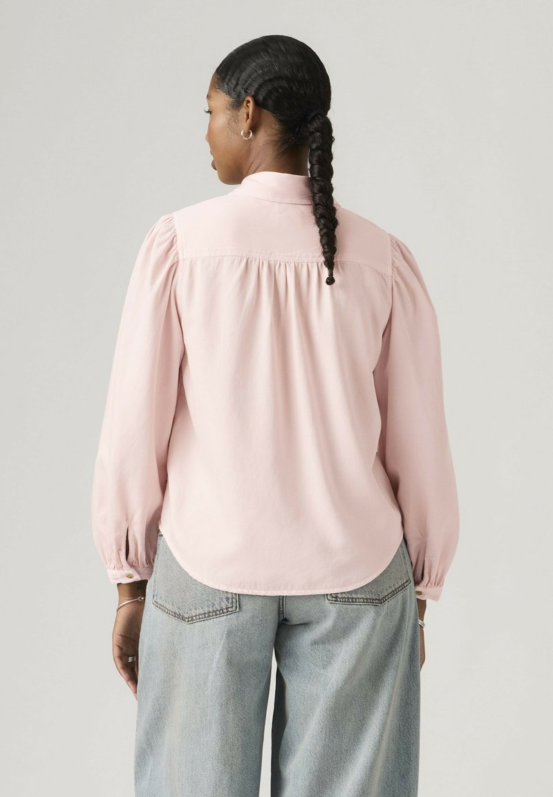 Femme aux cheveux tressés portant un chemisier rose clair à manches longues et un jean bleu clair, debout de dos contre un fond uni.