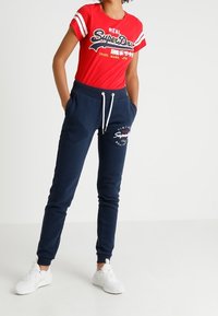 Röd T-shirt med vita ränder och logotyp, matchad med mörkblå joggers med dragsnöre och logotypdetalj, kompletterad med vita sneakers.