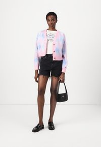 Cárdigan recortado de argyle en tonos rosa pastel y azul con botones, llevado sobre una camiseta gráfica blanca, pantalones cortos negros desgastados y mocasines negros, sosteniendo un bolso pequeño.
