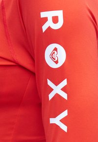 Κόκκινο πουκάμισο με μακριά μανίκια και λευκό κάθετο κείμενο "ROXY" και ένα λογότυπο σε σχήμα καρδιάς στο μανίκι κοντά στον αγκώνα.