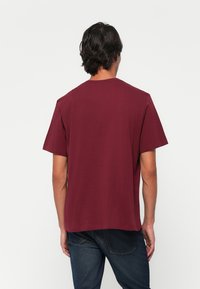 Man standing facing away wearing a plain burgundy short-sleeve t-shirt and dark blue jeans against a white background.