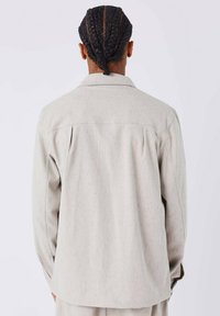 Giacca grigia chiara con texture, colletto a punta, maniche lunghe e vestibilità rilassata. Presenta un yoke posteriore e dettagli di fibre sottili.