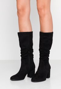 Svarta mocka knähöga stövlar med slouchy design, fyrkantig tå och 7,5 cm blockklack, med en sidodragkedja för enkel påtagning.