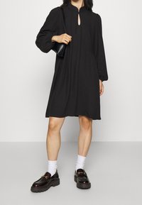 Svart klänning med halsringning i nyckelhålsdesign, långa puffärmar och lös passform. Matchas med kraftiga svarta loafers och vita ankelsockor.