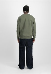 Felpa khaki con polsini a coste, collo a girocollo e dettagli nel pannello posteriore, abbinata a pantaloni in denim scuri a gamba larga con cuciture in evidenza.