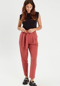 Pantalons taille haute plissés de couleur rouille avec une ceinture en tissu, associés à un haut noir ajusté sans manches et des mocassins noirs.