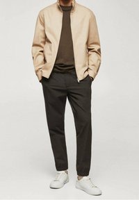 Jasnobeżowa kurtka na zamek błyskawiczny na ciemnobrązowym swetrze, w połączeniu z zwężanymi czarnymi spodniami i białymi trampkami, prezentująca nowoczesny, casualowy styl.