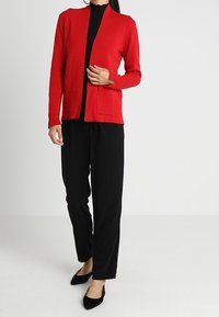 Cardigan rosso con apertura frontale, due tasche anteriori e maniche lunghe, abbinato a pantaloni neri e ballerine nere. Tessuto a maglia liscia.