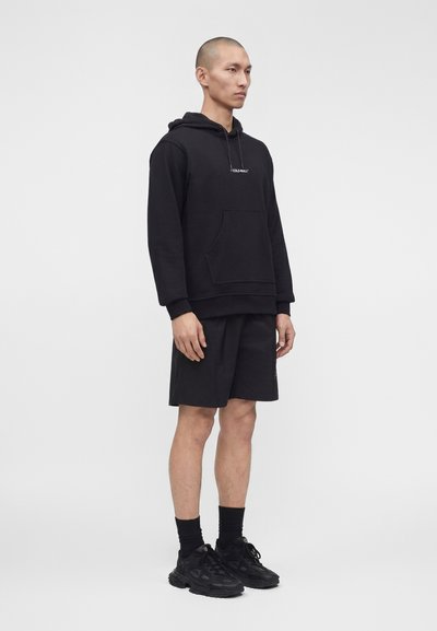 Άνδρας με μαύρο φούτερ με τσέπη μπροστά, μαύρα σορτς μέχρι τα γόνατα, μαύρες κάλτσες και μαύρα αθλητικά παπούτσια, που στέκεται μπροστά σε απλό λευκό φόντο.