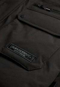 Czarna torba sportowa o gładkiej fakturze, z wyraźnym nadrukiem logo, kieszenią na zamek oraz płaskim panelem z przodu.