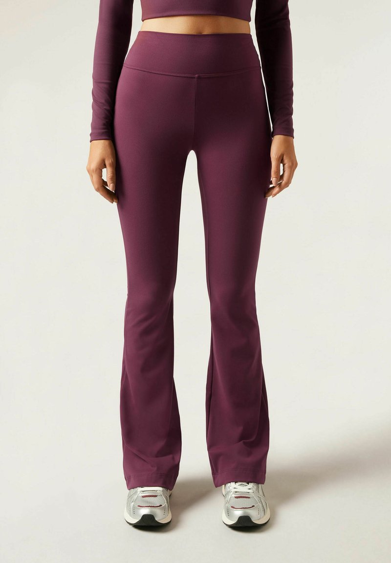 Flared workout leggings in dark purple, featuring a high waist and smooth texture. Paired with silver athletic shoes, showcasing subtle detailing.