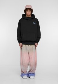 Sudadera negra con capucha y bolsillo frontal, combinada con pantalones de pierna ancha en un degradado pastel y zapatillas blancas, rematada con un sombrero de cubo rosa.