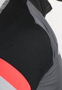 Camisa de rendimiento de manga corta con un cuerpo negro, acentos grises y una franja roja llamativa. Fabricada con una tela suave y elástica.