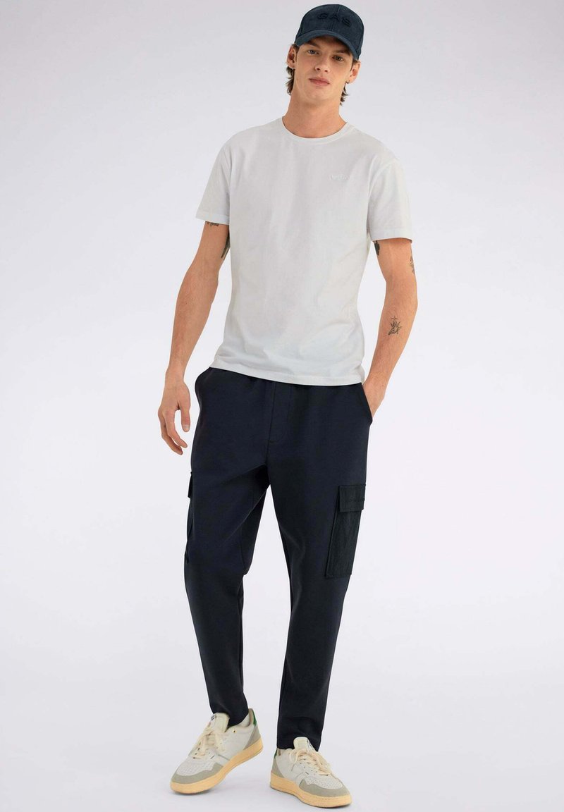 T-shirt grigio chiaro a maniche corte, pantaloni cargo blu navy con tasche laterali e sneakers beige con accenti verdi. Design semplice e funzionale.