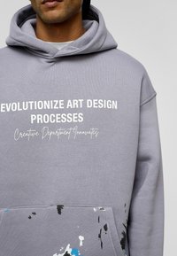 Grijze hoodie met een grote buidelzak aan de voorkant, witte tekst en een spetterpatroon in blauw en zwart aan de onderrand van de zak. Zachte stofstructuur.